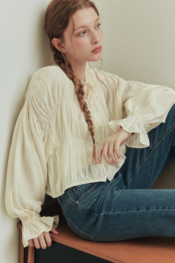 버니 셔링 블라우스 / 크림은 로맨틱한 무드를 연출하는 여성 블라우스입니다. 부드러운 레이온과 나일론 혼방 소재로 제작되어 봄, 여름 시즌에 시원하게 착용하기 좋습니다. 어깨, 가슴, 허리 라인에 셔링 디테일을 더해 여성스러운 실루엣을 강조하며, 리본넥 디자인과 긴 소매 기장으로 우아함을 더했습니다. 크롭 기장과 릴랙스핏으로 편안하면서도 세련된 데일리룩, 데이트룩, 카페룩, 모임룩을 완성해보세요. 반투명 소재와 작은 버튼 디테일, 드롭 숄더 디자인이 은은한 포인트를 더해줍니다.
